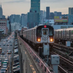 An elevated New York City Subway 7 train approaches on overhead tracks above a busy street filled with cars and buses.