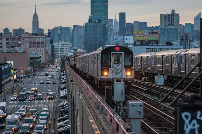 An elevated New York City Subway 7 train approaches on overhead tracks above a busy street filled with cars and buses.
