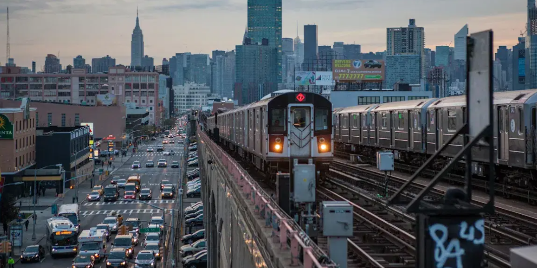 An elevated New York City Subway 7 train approaches on overhead tracks above a busy street filled with cars and buses.