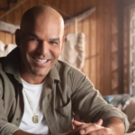 Puerto Rican actor and producer Amaury Nolasco smiles while seated indoors in a rustic wooden cabin, featured as part of a Discover Puerto Rico campaign.