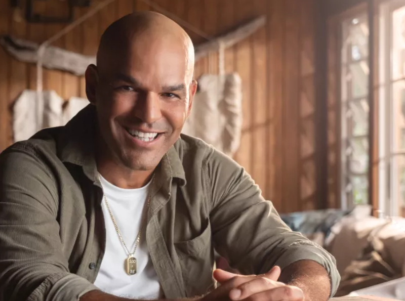 Puerto Rican actor and producer Amaury Nolasco smiles while seated indoors in a rustic wooden cabin, featured as part of a Discover Puerto Rico campaign.