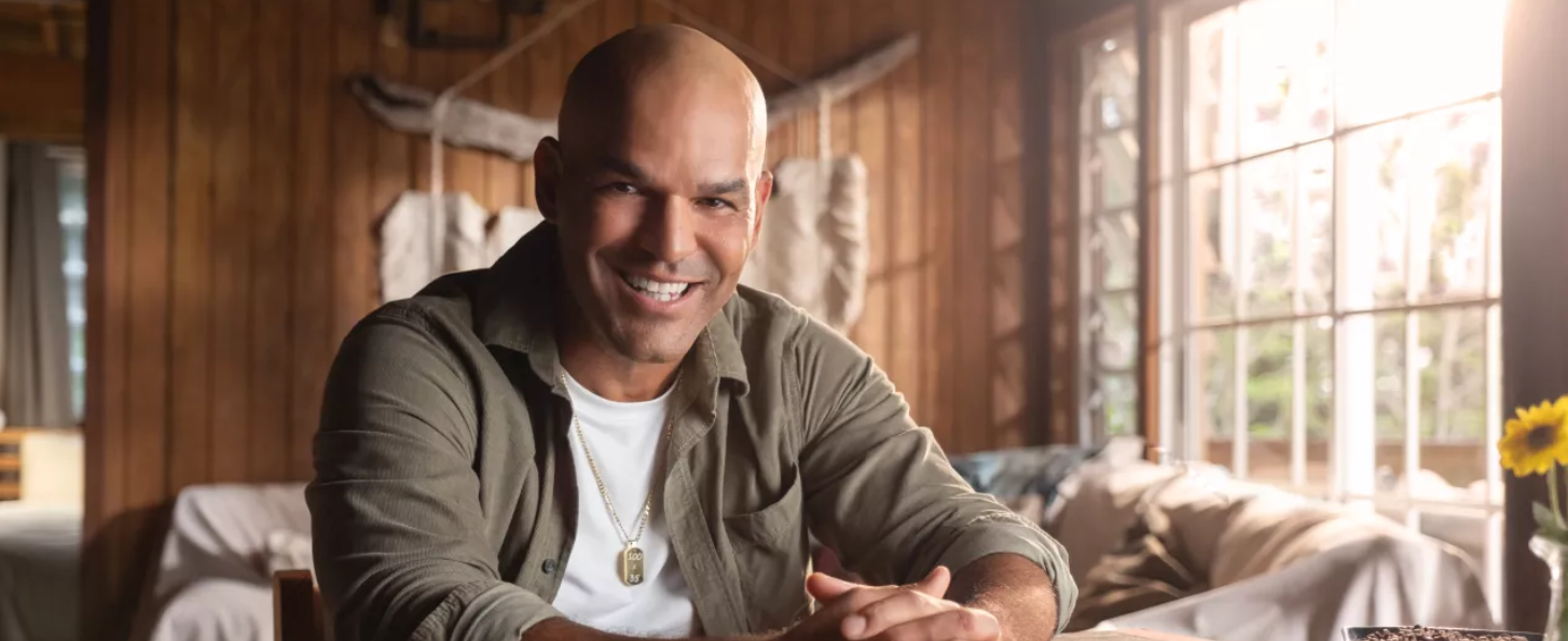Puerto Rican actor and producer Amaury Nolasco smiles while seated indoors in a rustic wooden cabin, featured as part of a Discover Puerto Rico campaign.