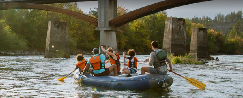 A family rafting on a river beneath a bridge, surrounded by trees and forested hills.