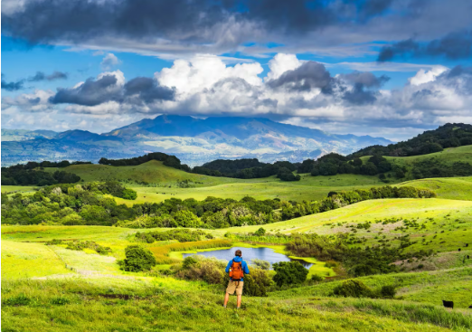 A hiker overlooking lush green hills, a small lake, and distant mountains beneath a partly cloudy sky.